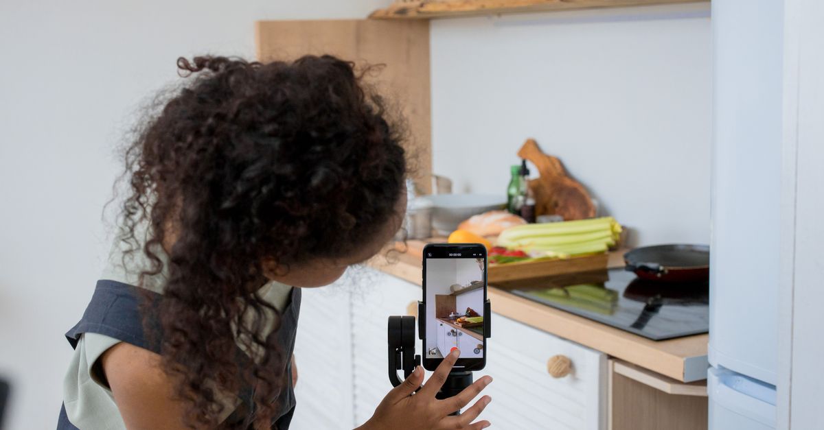 A woman with curly hair films a cooking video using a smartphone stabilizer in a modern kitchen.