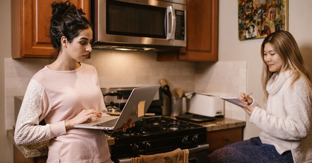Two women working from home in the kitchen with a laptop and notebook, focused and serious.
